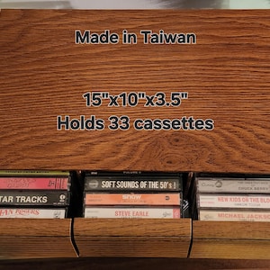 Puede incluir: Un mueble de almacenamiento de cintas de cassette de madera, hecho en Taiwán, con tres compartimentos. La unidad mide 38 cm x 25 cm x 9 cm y tiene capacidad para 33 casetes. La unidad está llena de casetes, con texto visible.