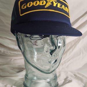 Op de afbeelding: Een marineblauwe baseballcap met een gele rechthoekige patch met het Goodyear-logo. De tekst "GOOD YEAR" is geel geborduurd. De cap heeft een gebogen klep en een mesh achterkant.
