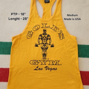 May include: Mustard yellow tank top with the text "GOLD'S GYM Las Vegas" and a graphic of a muscular man lifting weights. The tank top is a medium size, made in the USA, with a PTP of 18 inches and a length of 28 inches.