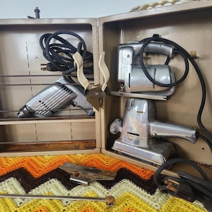Könnte beinhalten: Ein offener, vintage Werkzeugkoffer aus Metall mit zwei Elektrowerkzeugen und Zubehör. Der Koffer ist beige mit einem Verschluss und einem Griff. Die Werkzeuge sind silberfarben mit schwarzen Kabeln. Eine gelb, orange und braun gemusterte Decke befindet sich im Vordergrund.