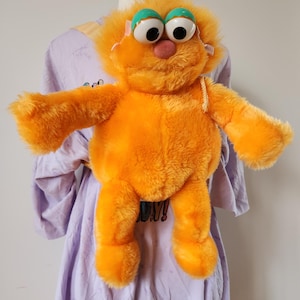 May include: An orange plush toy with large, cartoonish eyes and a pink nose. The toy has a furry texture and outstretched arms. The eyes are white with black pupils and teal eyelids. The toy is on a purple shirt.