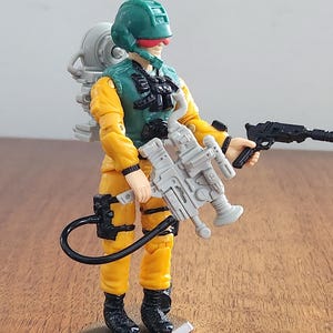 May include: A vintage action figure of a soldier in a yellow jumpsuit, green vest, and helmet. The figure is holding two toy guns, one gray and one black. It has a gray backpack and black boots. The figure is standing on a brown base.