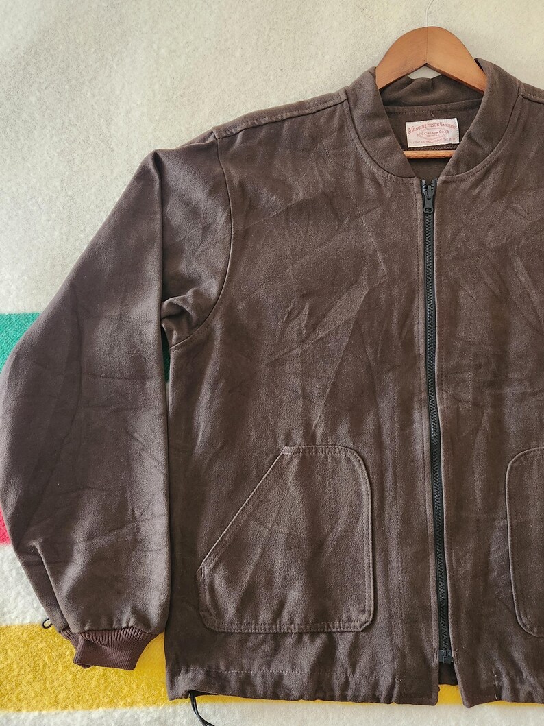 Peut inclure: Veste de bombardier marron avec une fermeture &eacute;clair noire et deux poches avant. La veste a un poignet c&ocirc;tel&eacute; et un col marron. La veste est suspendue &agrave; un cintre en bois.