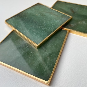 May include: Four square coasters with a green, marbled appearance. Each coaster has a gold-coloured metal border. The coasters are arranged at different angles, with three visible. The background is a plain white surface.