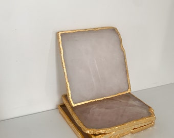 May include: A stack of four square coasters made of pale pink stone with gold-colored edges. The coasters are stacked on a white surface, with one coaster slightly elevated, showcasing the gold trim.