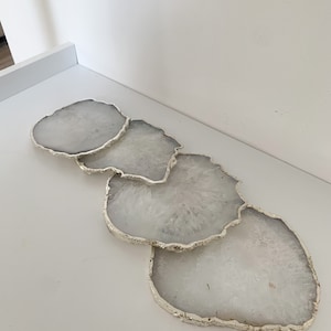 Puede incluir: Juego de cuatro posavasos de ágata blanca con borde metálico plateado. Los posavasos tienen una forma irregular y un aspecto natural y orgánico.