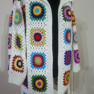May include: A white crocheted cardigan with a hood, featuring colorful circular patterns. The cardigan is open in the front and has long sleeves.