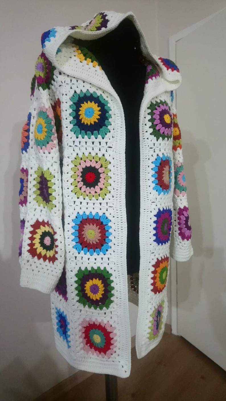 May include: A white crocheted cardigan with a hood, featuring colorful circular patterns. The cardigan is open in the front and has long sleeves.