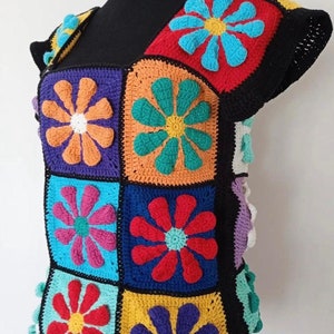 May include: A colorful crocheted top with a square pattern of flowers in various colors, including red, orange, yellow, green, blue, and purple. The top has short sleeves and a scoop neck.