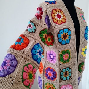 Op de afbeelding: Een beige gehaakt vest met kleurrijke vierkante motieven. De vierkanten zijn versierd met bloempatronen in roze, oranje, blauw, groen en paars. Het vest wordt getoond op een zwarte mannequin.