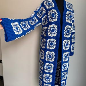 Könnte beinhalten: Ein langer, blauer Häkelcardigan mit einem weißen und blauen geometrischen Muster. Der Cardigan hat einen V-Ausschnitt und lange Ärmel.