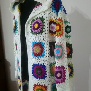 May include: A white crocheted cardigan with a hood, featuring colorful granny square motifs. The cardigan has a long, open front and long sleeves.