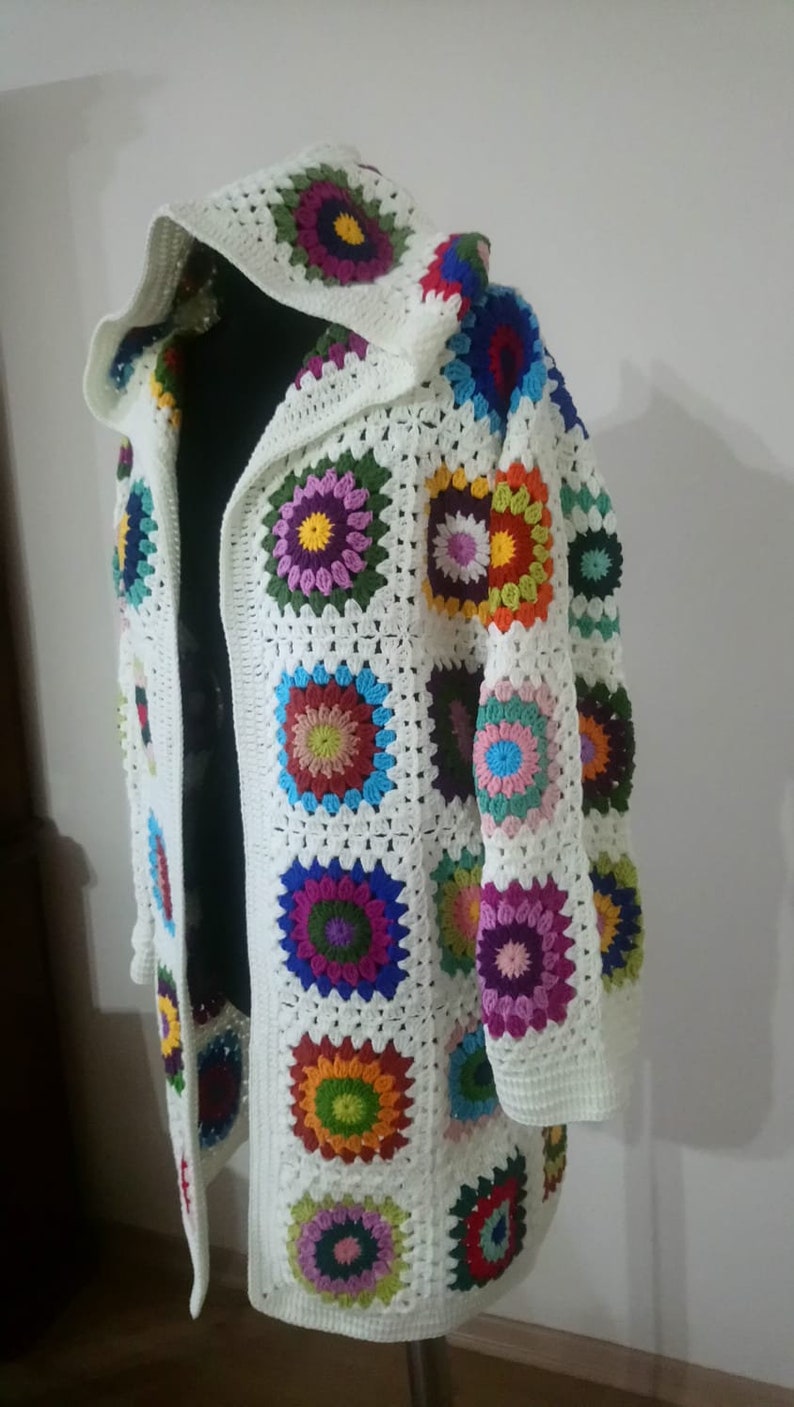 May include: A white crocheted cardigan with a hood, featuring colorful granny square motifs. The cardigan has a long, open front and long sleeves.