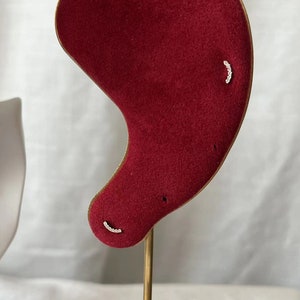 May include: A pair of small, curved earrings with a row of tiny white stones set in gold metal. The earrings are displayed on a red velvet ear model.