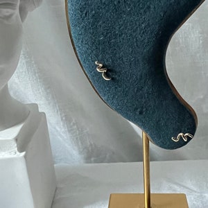 May include: Gold-colored stud earrings in a minimalist, abstract design, displayed on a teal velvet earring holder. The earrings have a delicate, wavy shape. The holder is mounted on a gold-colored stand with a square base.