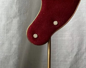 Puede incluir: Un expositor de joyas de oro con un busto de terciopelo rojo. Dos pequeños pendientes redondos se muestran en el busto.