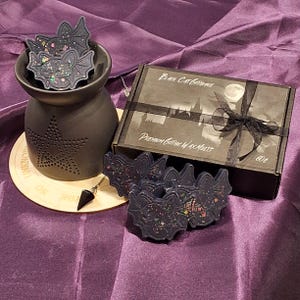 May include: A black wax melt warmer with a star design and a black bat-shaped wax melt. The wax melt is decorated with glitter. The warmer is on a wooden base with a black pendulum. The wax melts are packaged in a black box with a black ribbon and the text "Black Cat Gothique Premium Gothic Wax Melts".