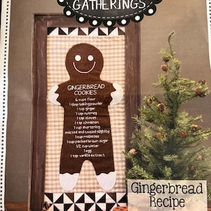 May include: A framed fabric wall hanging featuring a brown gingerbread person applique on a beige gingham background. The gingerbread person has white icing details and text listing a gingerbread cookie recipe. The frame is dark wood with black and white geometric trim.