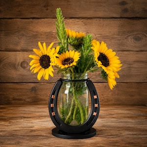 May include: A clear glass mason jar vase held within a black metal horseshoe frame featuring small copper-colored square accents. The jar contains a bouquet of bright yellow sunflowers with dark brown centers and green stems and leaves.