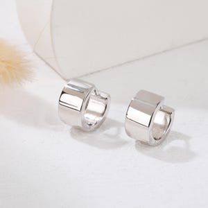May include: A pair of silver hoop earrings with a flat, rectangular design. The earrings are simple and modern.