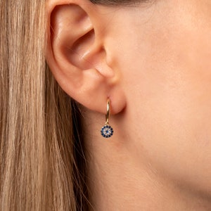 May include: Gold hoop earrings with a blue and white evil eye charm dangling from the bottom.
