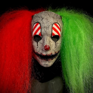 Puede incluir: Una máscara de payaso espeluznante con pelo rojo y verde. La máscara tiene una cara blanca agrietada con ojos rayados rojos y blancos y una nariz roja. La máscara tiene una sonrisa siniestra.