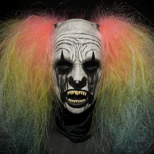 Puede incluir: Una máscara de payaso blanca con maquillaje negro y una amplia sonrisa con dientes. La máscara tiene una lágrima negra que le cae por cada mejilla. La máscara está rodeada de una peluca arcoíris con mechones rosas, amarillos y azules.