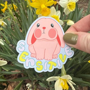 May include: A pink sticker featuring a cartoon bunny with rosy cheeks and the word "SENSITIVE" in yellow letters. The sticker is held in front of a backdrop of yellow and white daffodils.