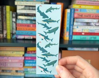 May include: A blue and green bookmark with a pattern of sharks swimming in a light blue ocean. The sharks are in various shades of blue and green, with white bellies and black eyes. The bookmark is rectangular and has a white border.
