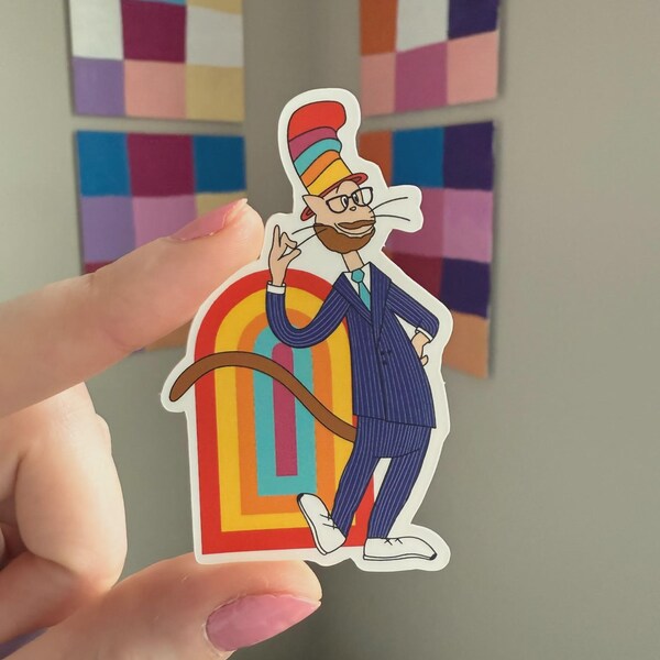 Op de afbeelding: Een kleurrijke sticker met een cartoon kattenpersonage in een krijtstreep pak, bril en een gestreepte hoed. De kat staat voor een regenboogboog. De sticker wordt door een hand omhoog gehouden.