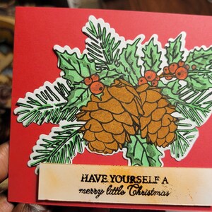 May include: A red Christmas card featuring a green holly and pine branch design with two brown pine cones. The card has a cream-coloured tag with the text "HAVE YOURSELF A merry little Christmas".
