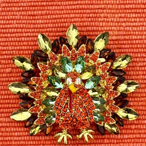 May include: A jeweled turkey brooch with a vibrant display of colors. The brooch features a body of orange gems, a green and gold tail, and a red beak. The brooch is set against an orange textured background.