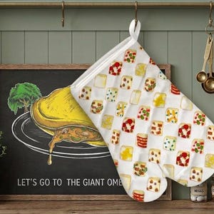 May include: A white oven mitt featuring a repeating pattern of various small, colorful food illustrations, including slices of bread with toppings, hanging from a hook. The mitt has a white fabric loop at the top for convenient storage.