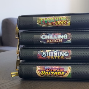 Op de afbeelding: Vier zwarte mappen met gouden ritsen gestapeld op elkaar. Elke map heeft een kleurrijke sticker met de naam van een Pokémon Trading Card Game-uitbreiding. De uitbreidingen zijn "Evolving Skies", "Chilling Reign", "Shining Fates" en "Vivid Voltage".