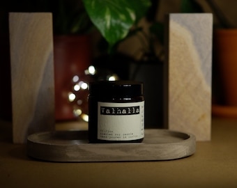 May include: A dark amber glass candle jar with a black lid and a white rectangular label. The label features the text Valhalla, 50/30h, scented soy candle, and hand-poured in Dublin. The jar sits on a smooth, oval-shaped gray concrete tray.