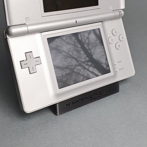 Könnte beinhalten: Eine silberne Nintendo DS Lite Handheld-Spielkonsole wird auf einem schwarzen Ständer präsentiert. Der Ständer trägt den Text "For Nintendo DS Lite".