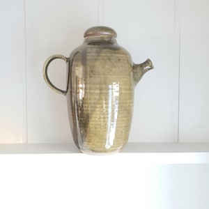 Puede incluir: Una jarra de cerámica con un esmalte verde claro. La jarra tiene un cuerpo redondeado y un pico en el lateral. La jarra está sobre una repisa blanca.