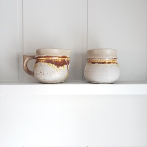 May include: Two ceramic mugs with a brown and white glaze. The mugs have a speckled texture and a rounded shape.