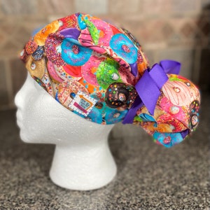May include: A patterned scrub cap with a design of colourful donuts in shades of pink, blue, and brown, with sprinkles. The cap has a purple bow and a tag that reads "Mom Aunt & Betty Designs".