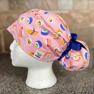 May include: A pink surgical scrub cap featuring a repeating pattern of sushi rolls in various colors. The cap has a dark blue ribbon and a small tag with text. The cap is designed to be worn during medical procedures.