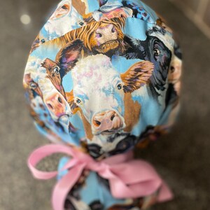 May include: A light blue scrub cap featuring a repeating pattern of cow faces in various colors. The cap is tied with a pink ribbon. The cow faces are in shades of brown, black, white, and tan.
