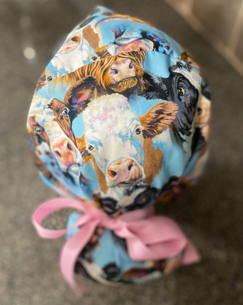 May include: A light blue scrub cap featuring a repeating pattern of cow faces in various colors. The cap is tied with a pink ribbon. The cow faces are in shades of brown, black, white, and tan.