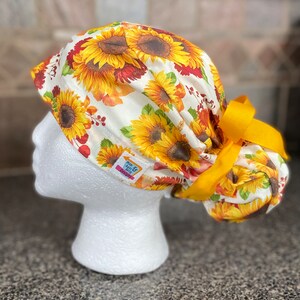 Peut inclure: Un bonnet de soin blanc orné d'un motif de tournesols aux couleurs vives : jaune, orange et marron. Le bonnet est orné d'un ruban jaune et d'une petite étiquette avec l'inscription "Miss April & Britt Design".