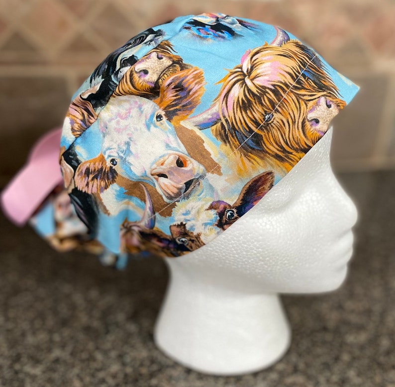 May include: A light blue scrub cap featuring a repeating pattern of watercolor-style cow faces in various colors. The cap has a pink bow on the back. The cow faces include white, brown, and black cows.