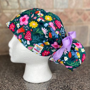 May include: A dark green scrub cap with a colorful floral pattern featuring pink, yellow, and white flowers, red mushrooms, and butterflies. The cap has a light purple ribbon and a small tag that reads "Mom & Aunt Busy Designs."