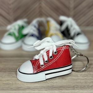 May include: A red miniature canvas sneaker keyring with white laces and a white toe cap. The keyring has a black and white sole and a silver metal ring. Other colourful sneakers are blurred in the background.