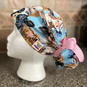 May include: A light blue scrub cap featuring a cow print with various cow faces. The cap has a pink bow on the side and a small tag that reads "Mum & Aunt Gitty Designs".
