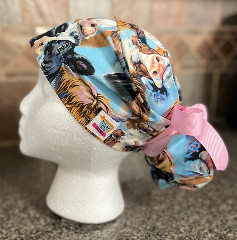 May include: A light blue scrub cap featuring a cow print with various cow faces. The cap has a pink bow on the side and a small tag that reads "Mum & Aunt Gitty Designs".