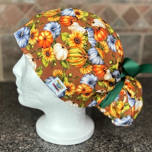 Peut inclure: Une charlotte de bloc opératoire sur fond marron avec un motif de citrouilles orange, blanches et bleues, de tournesols jaunes et de feuilles vertes. Elle possède un ruban vert à l'arrière et une petite étiquette blanche Mom & Aunt E Designs.