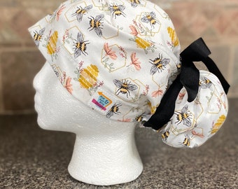 May include: White scrub cap with a bee and honeycomb pattern. The cap features a black bow and a tag with the text "Mom & Light Etsy Designs". The design includes bees, honeycombs, and floral elements in yellow, pink, and gray.
